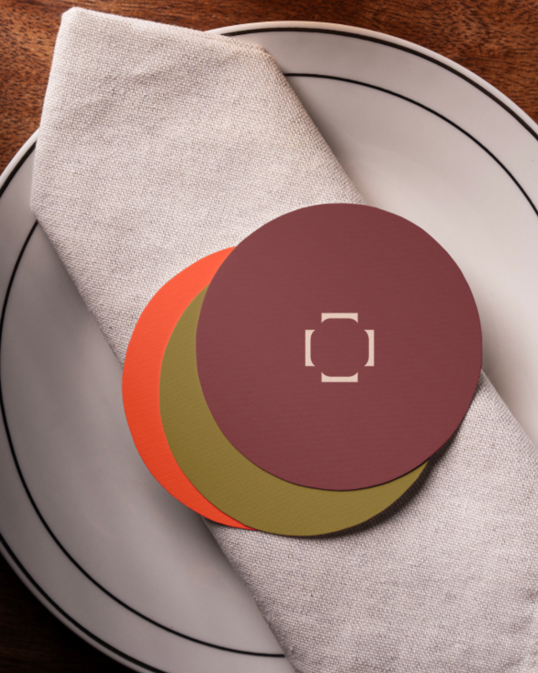 Dessous de verres de couleurs bordeaux, olive et orange du restaurant Table présentée en pastilles rondes sur une assiette et une serviette en lin