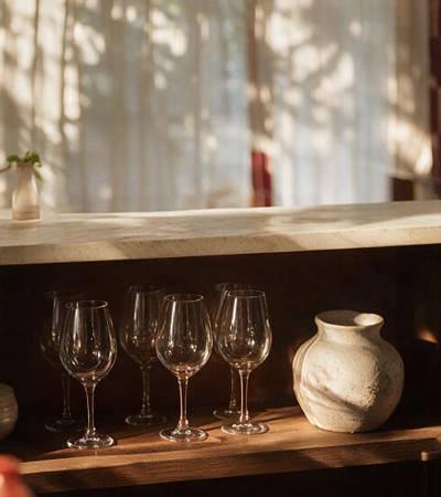 Verres à vin alignés sur une étagère en bois avec vase en céramique brute, lumière tamisée à travers un voilage
