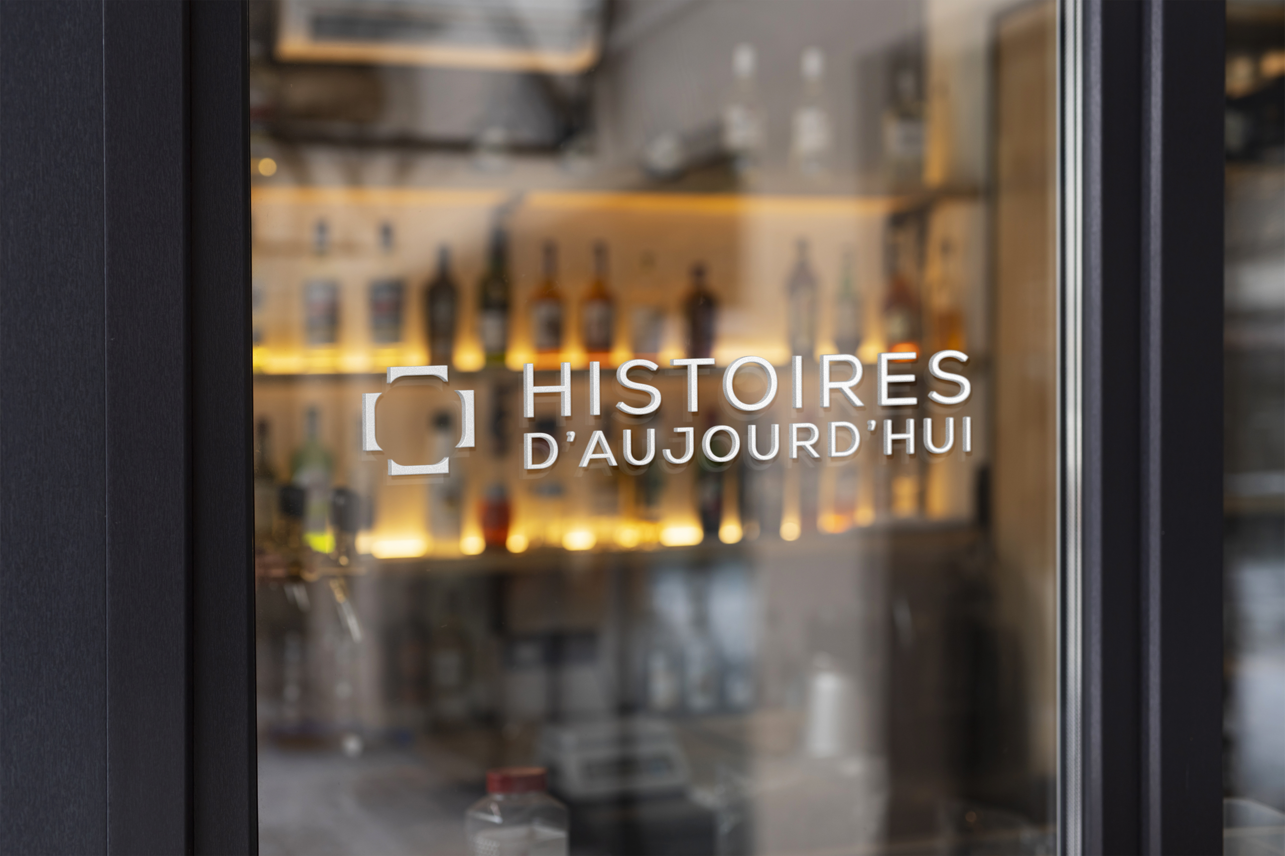 Logo "Histoires d'aujourd'hui" gravé sur la porte vitrée du restaurant Table, bar et bouteilles en arrière-plan flouté